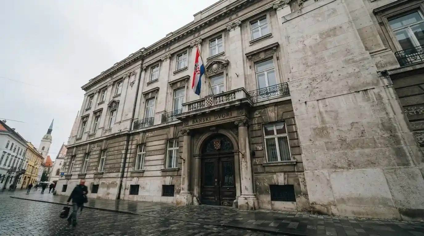 Pročelje zagrebačke ministarske zgrade od sivog kamena s visokim vratima i zastavom, oblačno nebo iznad.